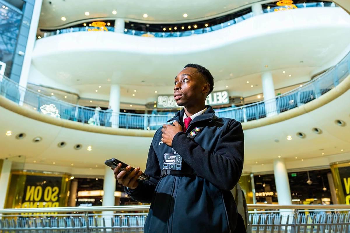 A young black security guard holding his radio, with levels of a shopping centre seen behind him