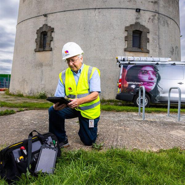 A man in Mitie hard hat and high vis kneeling in front of a concrete building and using a digital tablet